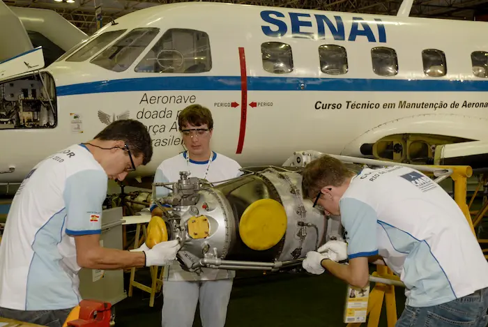 Alunos do SENAI trabalham em um motor de aeronave em frente a um avião com a inscrição 'Curso Técnico em Manutenção de Aeronaves