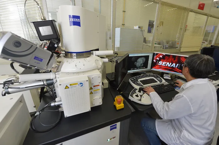 Técnico do SENAI opera um microscópio eletrônico em um laboratório, analisando imagens em dois monitores à sua frente.