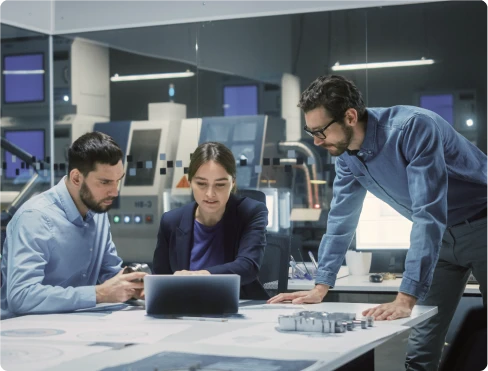 Imagem de três profissionais fazendo reunião em um ambiente tecnológico. Estão em torno de uma mesa com laptops e papéis técnicos.